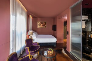 Habitación de hotel con cama, un sillón morado, una mesita con fruta y un cuadro en la pared.