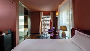 A hotel room with a bed, a purple armchair, a small table with a lamp, and a view of the city through large windows. - Casa Suzanna (Tel Aviv)