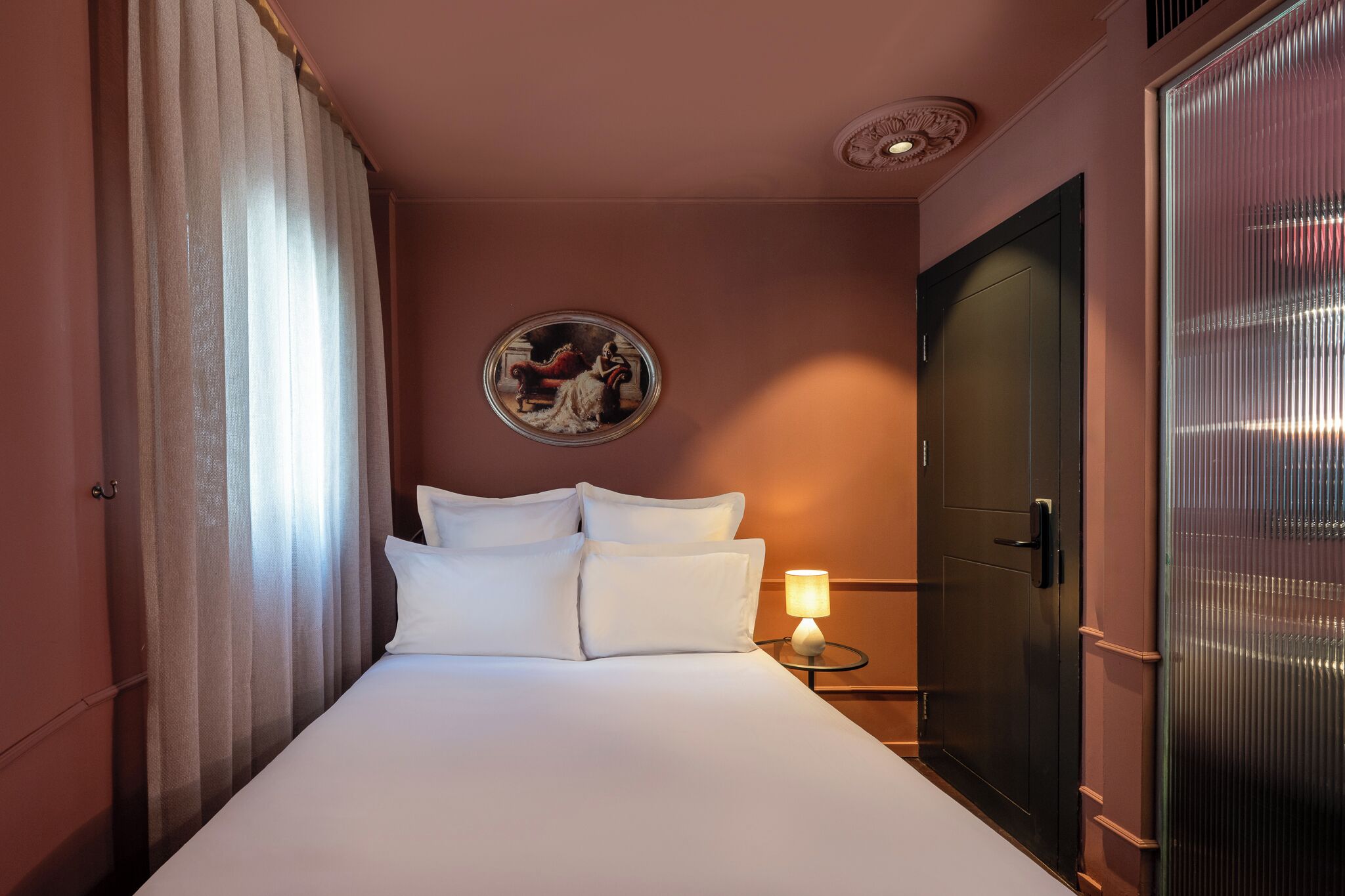 A hotel room with a bed, a nightstand with a lamp, a door, and a wall-mounted painting.