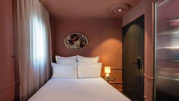 A hotel room with a bed, a nightstand with a lamp, a door, and a wall-mounted painting.