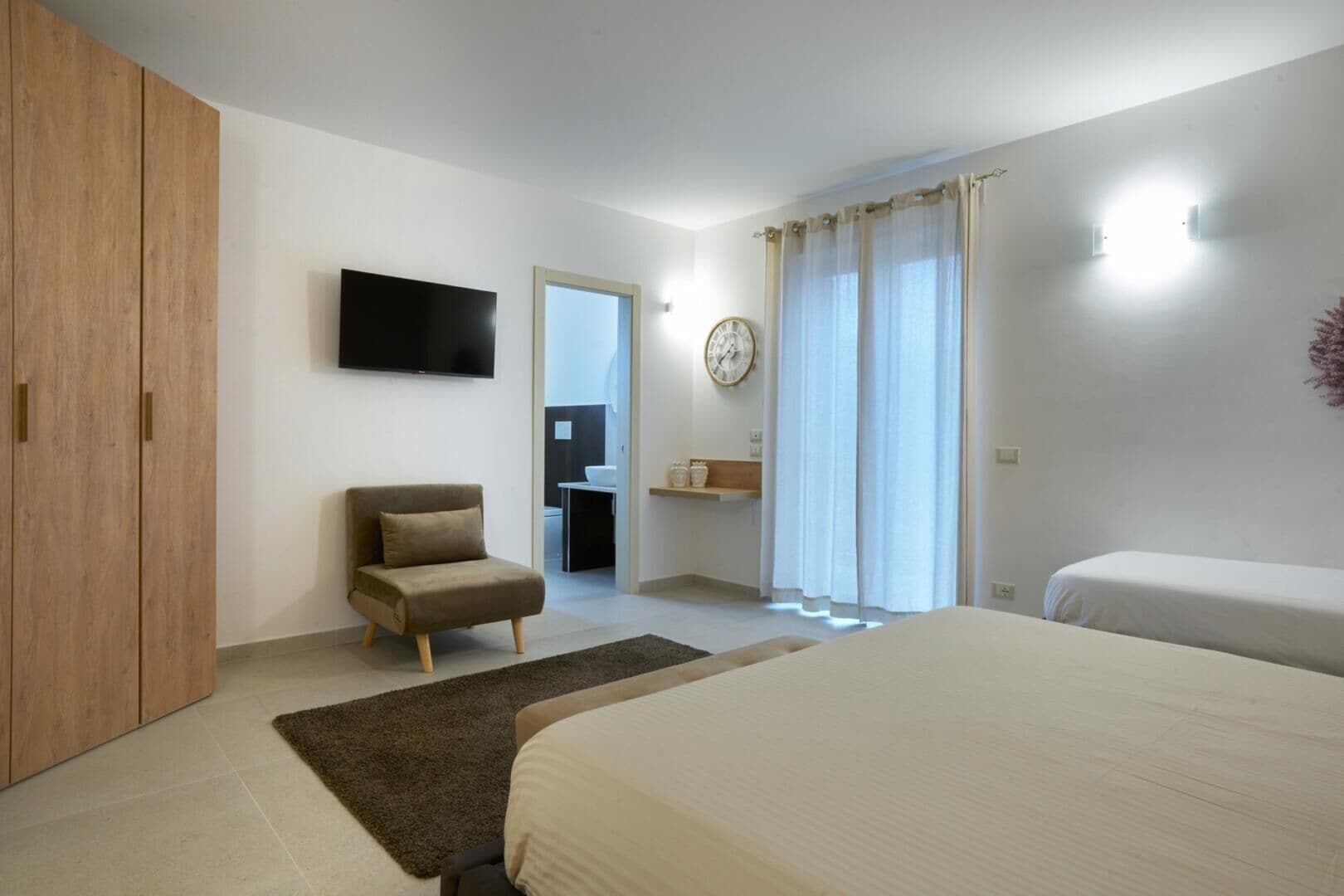 Quarto com cama, cadeira, guarda-roupa, televisão, relógio e banheiro visível através de uma porta aberta.