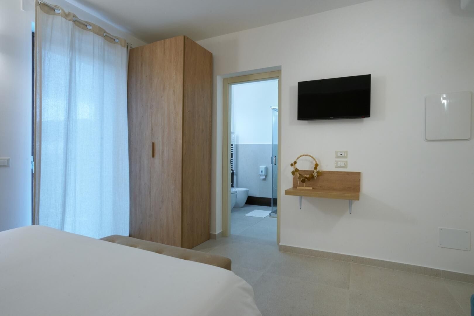 Una habitación de hotel con cama, un armario de madera, un televisor en la pared y un pequeño estante de madera con un objeto decorativo.