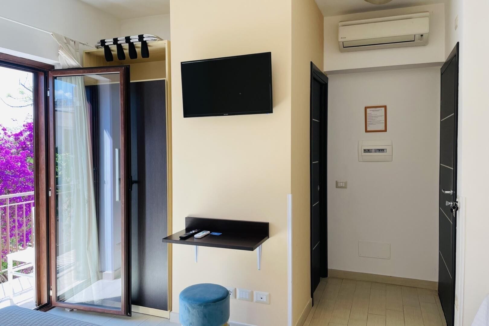 Una habitación moderna con una puerta de cristal corredera que da a un balcón, un televisor de pantalla plana en la pared, un estante negro pequeño y un otoman azul.