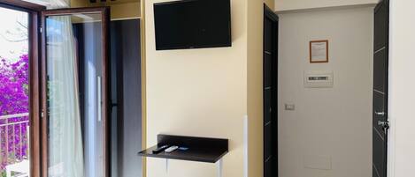 Una habitación moderna con una puerta de cristal corredera que da a un balcón, un televisor de pantalla plana en la pared, un estante negro pequeño y un otoman azul.