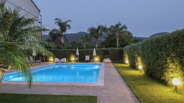 A rectangular swimming pool with a tiled edge, surrounded by a paved walkway. There are lounge chairs and umbrellas along the poolside. The area is well-lit with ground lights, and there are palm trees and a hedge in the background. - Il Girasole Capo d'Orlando (Capo d'Orlando)