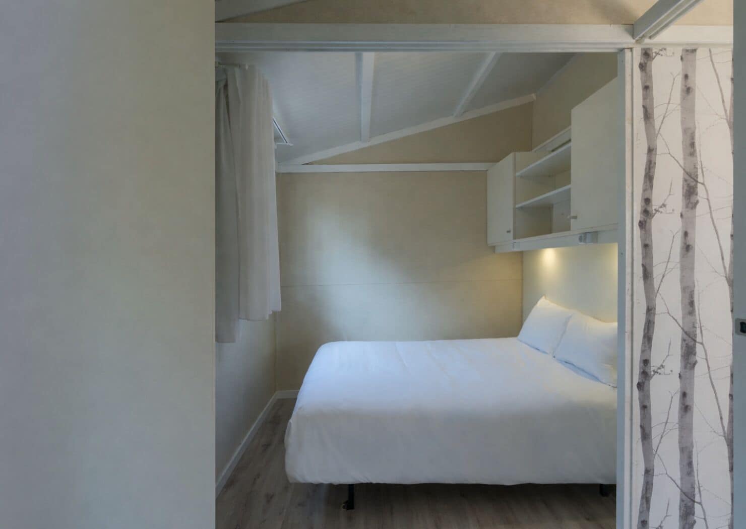 Una piccola camera con un letto singolo, pavimento in legno, un letto bianco e uno scaffale a parete.