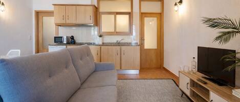 A modern living room with a grey sofa, a TV, and a kitchen area with wooden cabinets and a microwave.