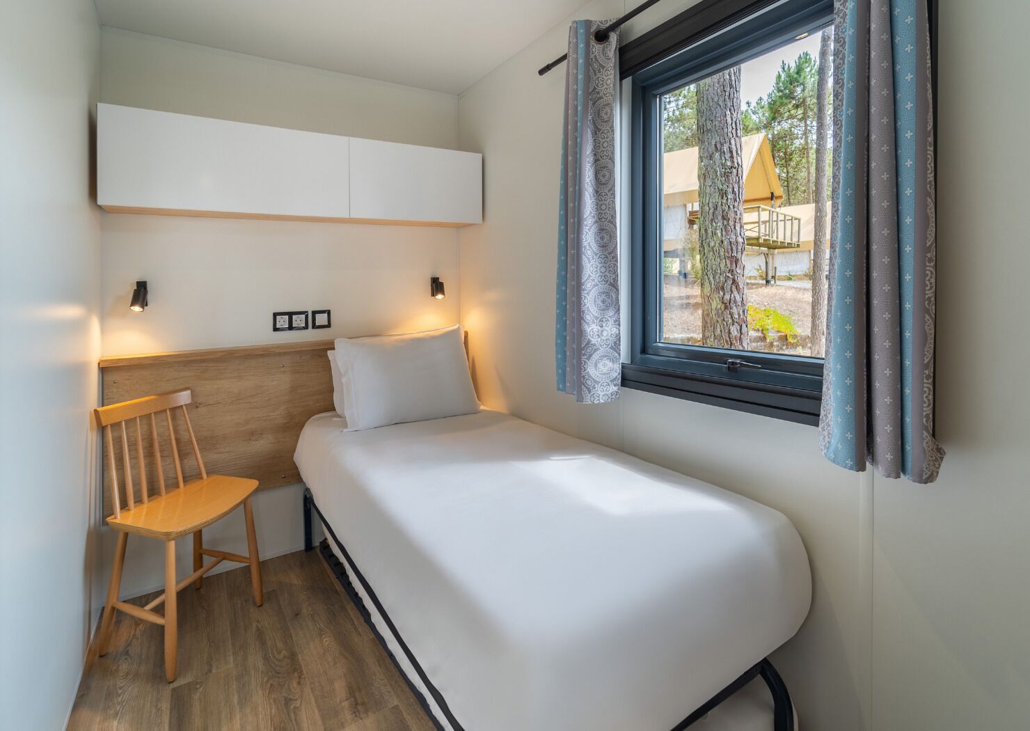Une petite chambre avec un lit simple, une tête de lit en bois, une chaise, une table de chevet et une fenêtre avec des rideaux.
