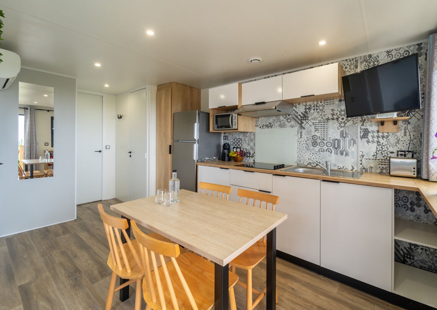 A modern kitchen with a dining area, wooden chairs, a stainless steel refrigerator, a microwave, and a flat-screen TV mounted above the counter.