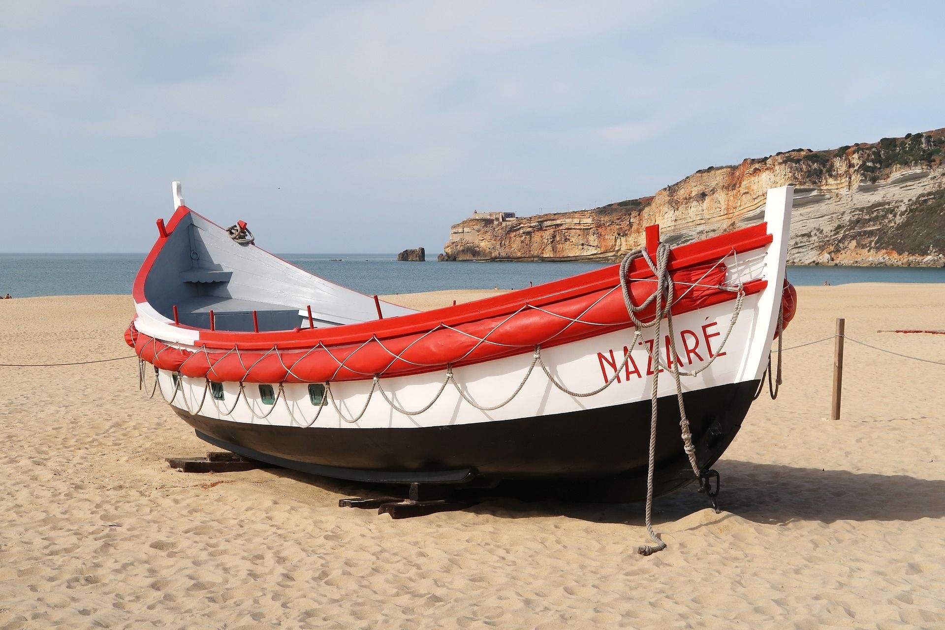 Un bateau nommé « NAZARE » est échoué sur une plage de sable, avec des falaises en arrière-plan.