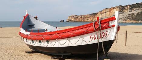 Un bateau nommé « NAZARE » est échoué sur une plage de sable, avec des falaises en arrière-plan.