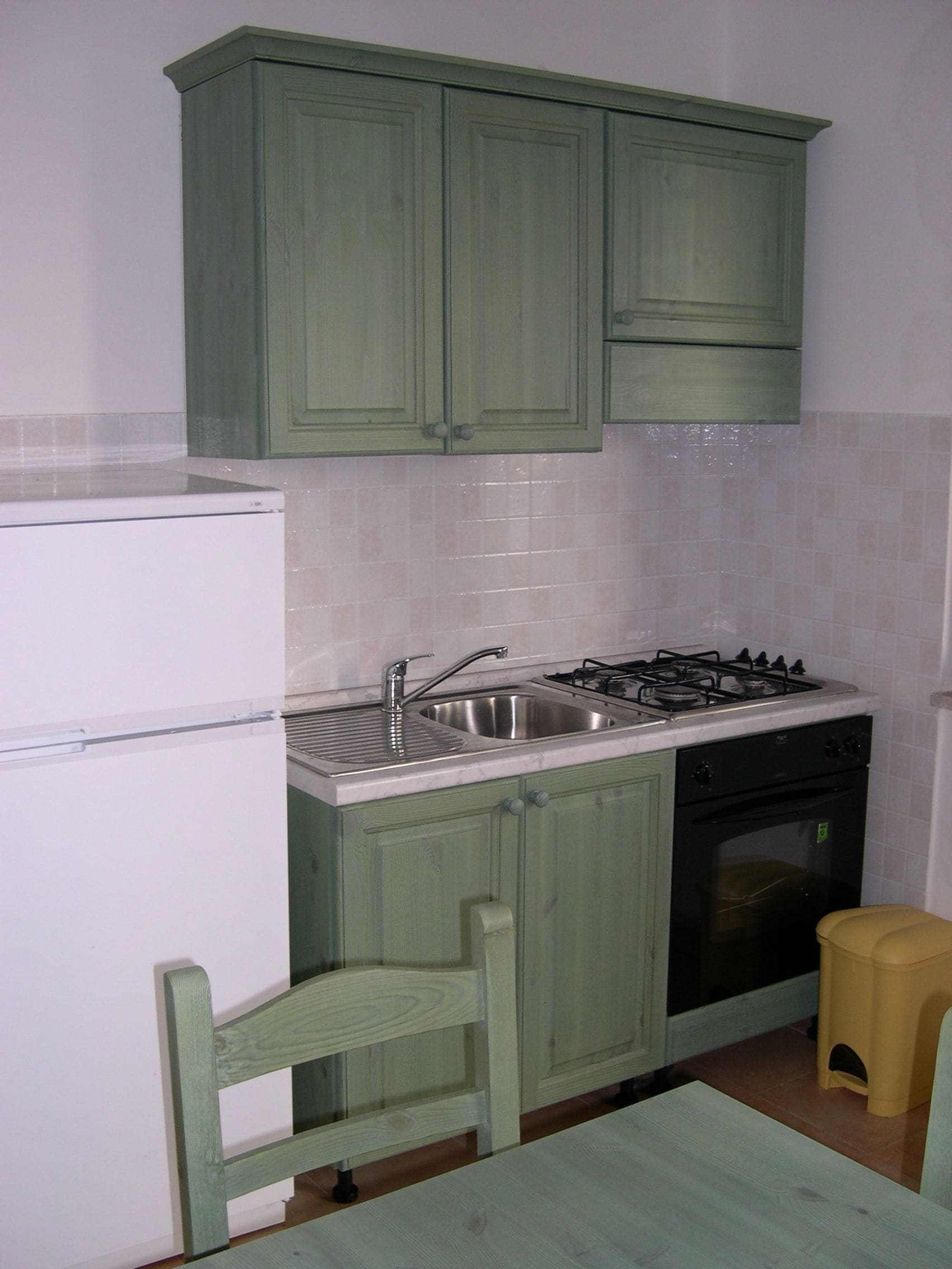 A compact kitchen with green cabinets, a white refrigerator, a sink, a stove, and a small stool.