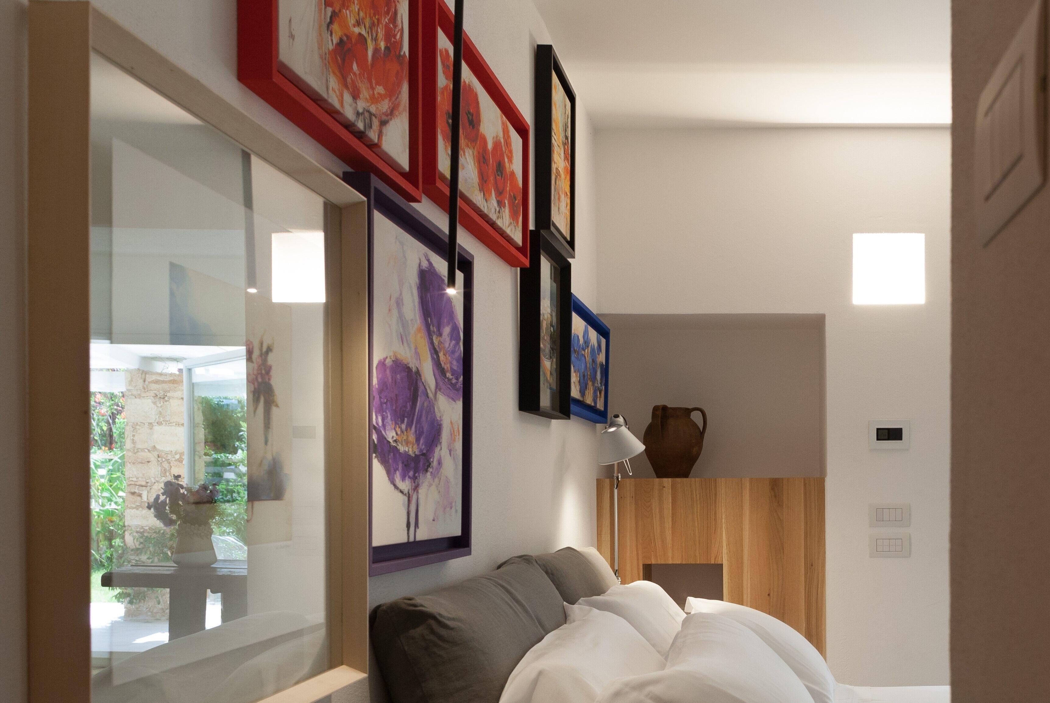 A modern bedroom with a bed, a nightstand, and a vase. There are framed artworks on the wall and a window with a view of greenery.
