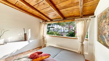 Una habitación con cama, ventana con cortinas, vistas a árboles y flores, y techo de madera.