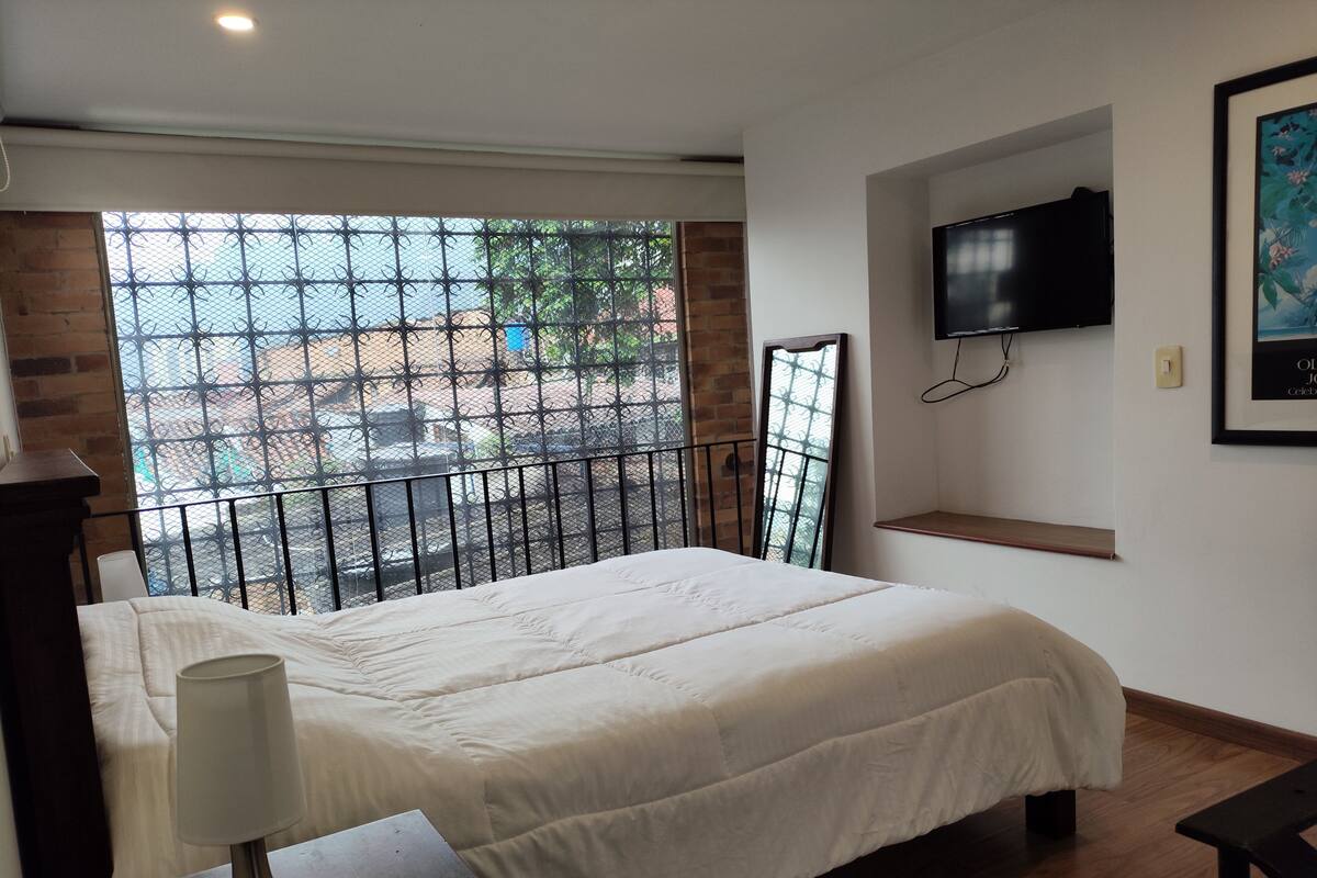 Une chambre à coucher avec un lit, un téléviseur fixé au mur, une fenêtre avec un moustiquaire en métal et un mur en briques.
