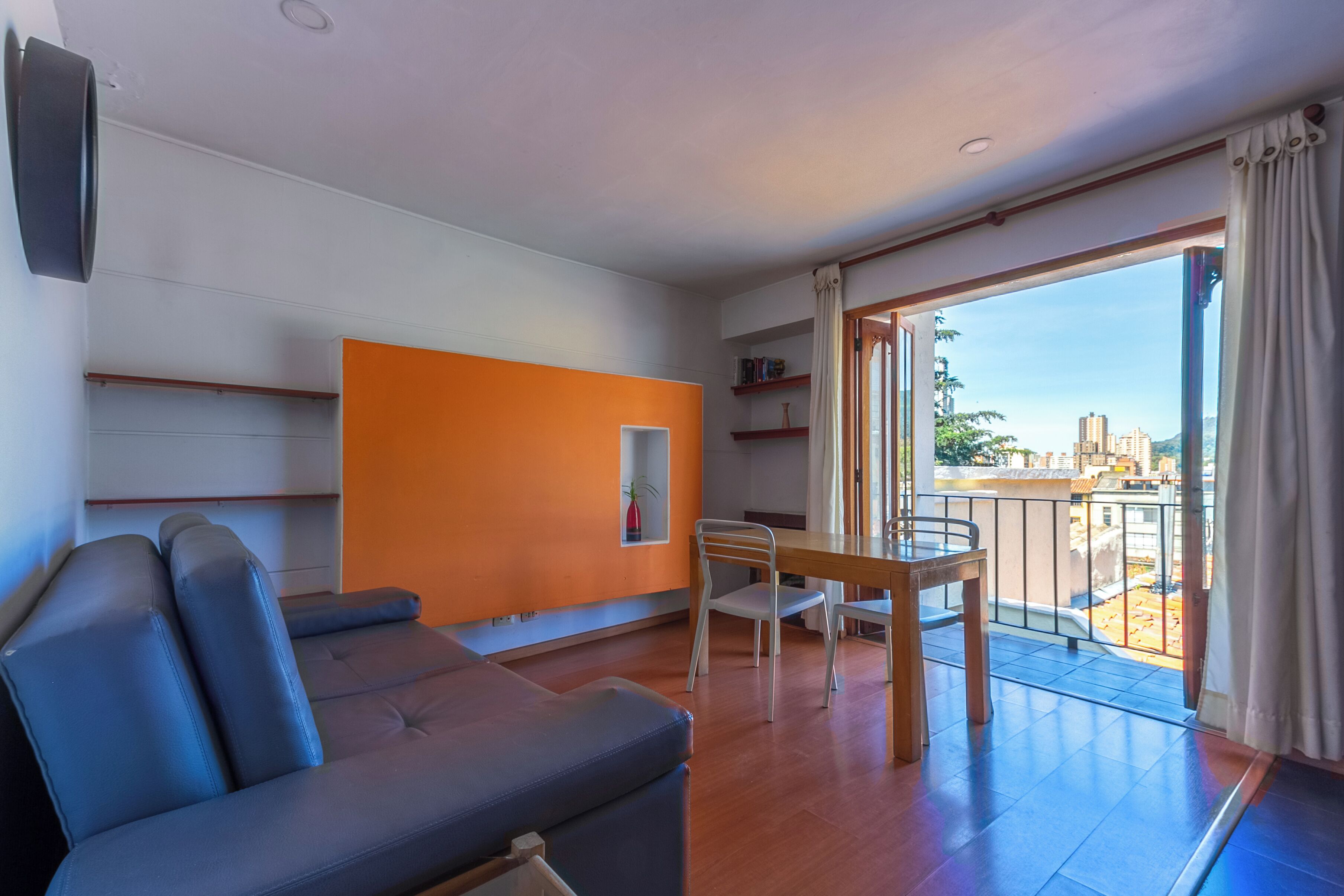 A modern living room with a sofa, a dining table, and chairs. There is a balcony with a view of the city.