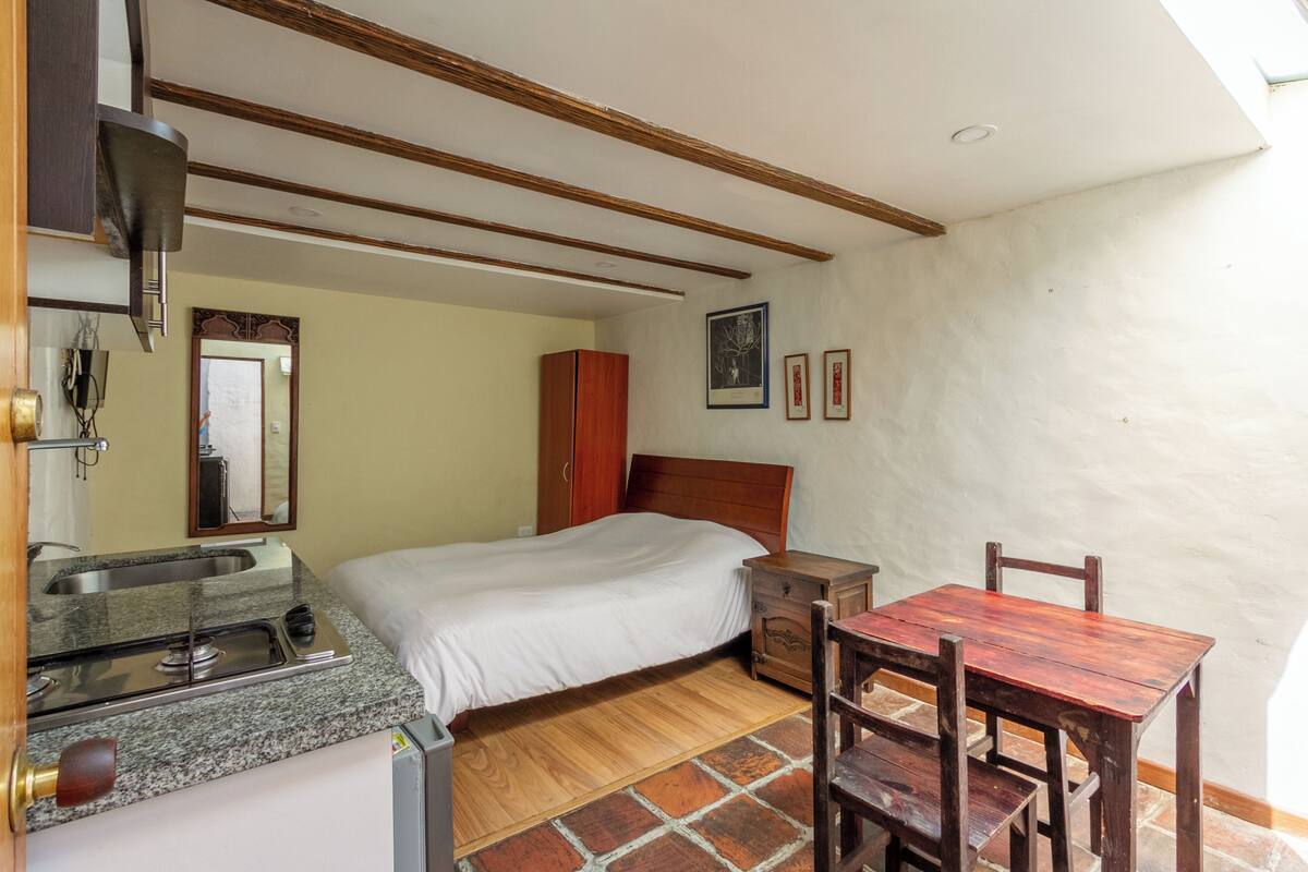 Une chambre à coucher comprenant un lit, une tête de lit en bois, une table de chevet, une table en bois avec des chaises, un petit coin cuisine avec un évier et un sol carrelé.