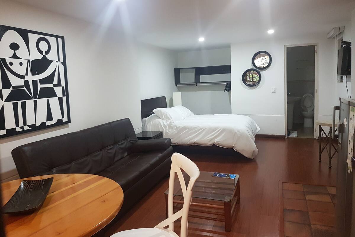 Une chambre d’hôtel moderne avec un canapé en cuir noir, une table à manger en bois, un lit avec une literie blanche, une petite télévision et un miroir fixé au mur.