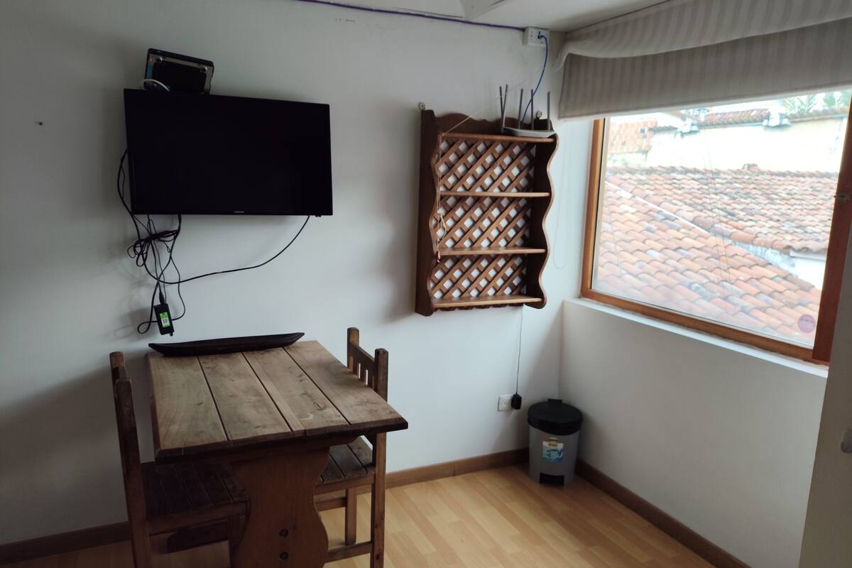 Une petite pièce avec une table et des chaises en bois, un téléviseur fixé au mur, une fenêtre donnant sur un toit recouvert de tuiles et une étagère murale.