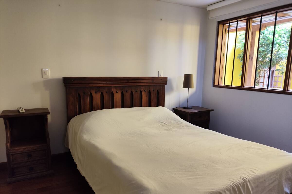 Une chambre à coucher avec une tête de lit en bois, une table de chevet avec une lampe et une fenêtre avec des stores.
