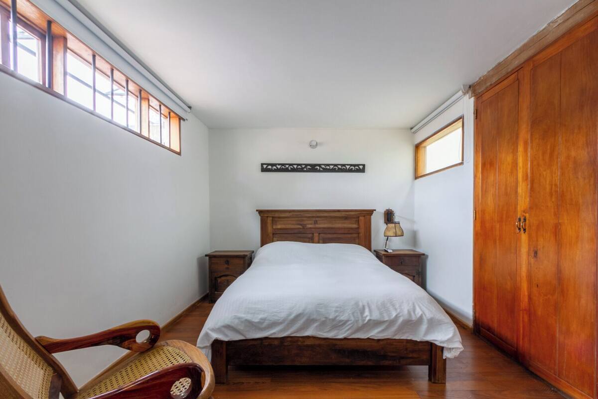 Une chambre à coucher avec un lit en bois, une table de chevet, une armoire en bois, une chaise en osier et une fenêtre avec des stores.