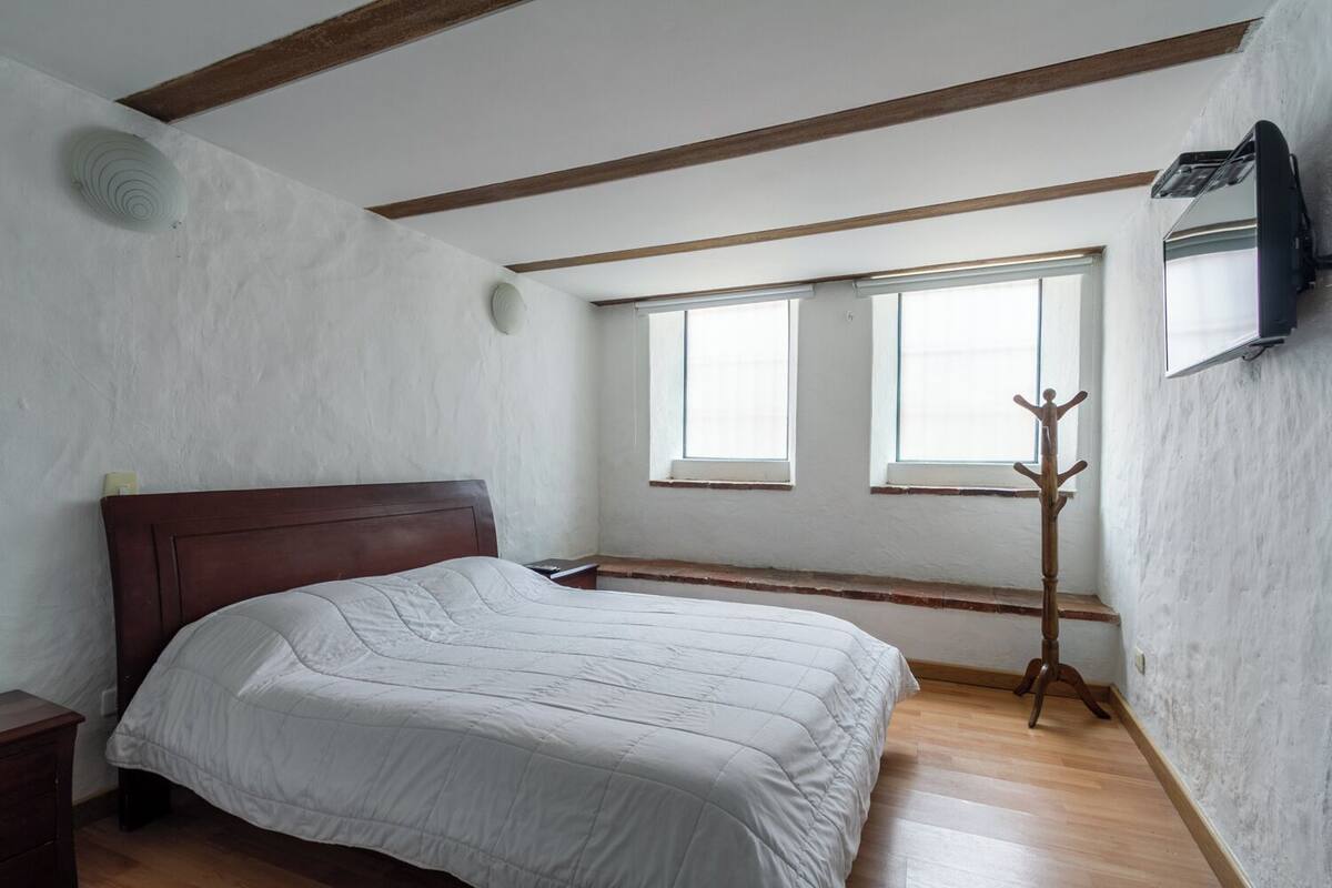 Une chambre à coucher avec un lit en bois, deux fenêtres, un porte-manteau et un climatiseur fixé au mur.