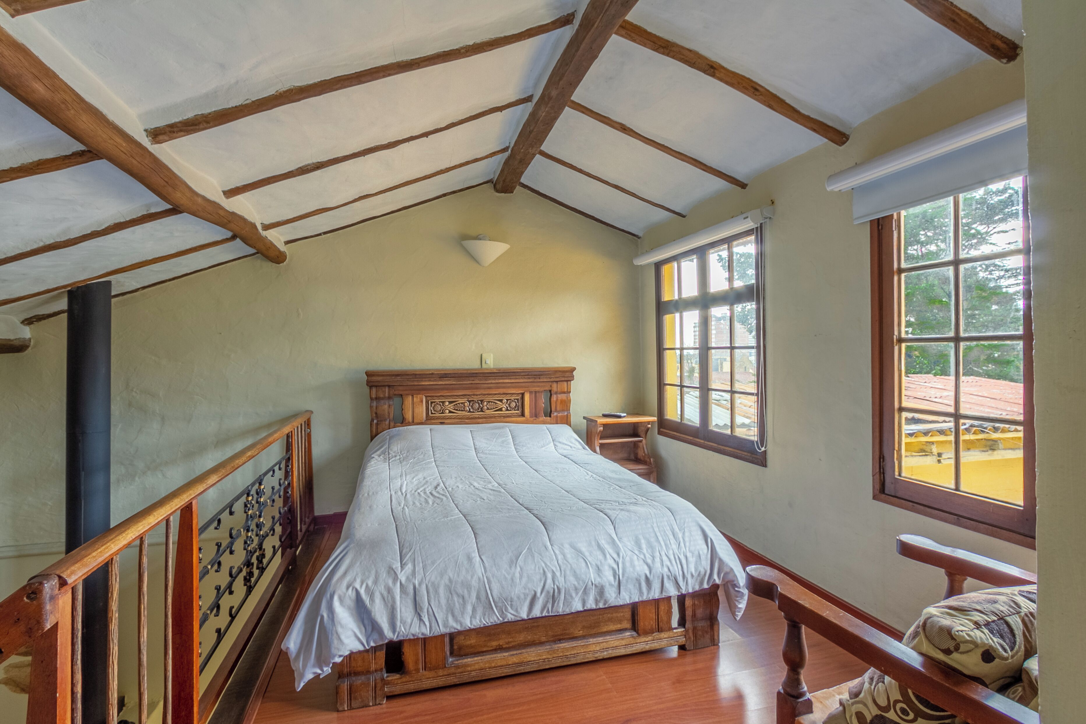 Un dormitorio con cama de madera, ventana con vista a la vegetación y techo inclinado con vigas de madera.