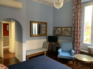 A room with a bed, a blue armchair, a small table, a television, and a mirror. There are floral curtains and a window with a view. - Relais Teatro Argentina (Rome)