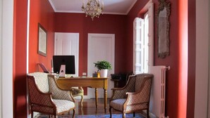 Quarto com paredes vermelhas, um candelabro, uma escrivaninha de madeira, duas cadeiras estampadas, um radiador e um espelho grande.