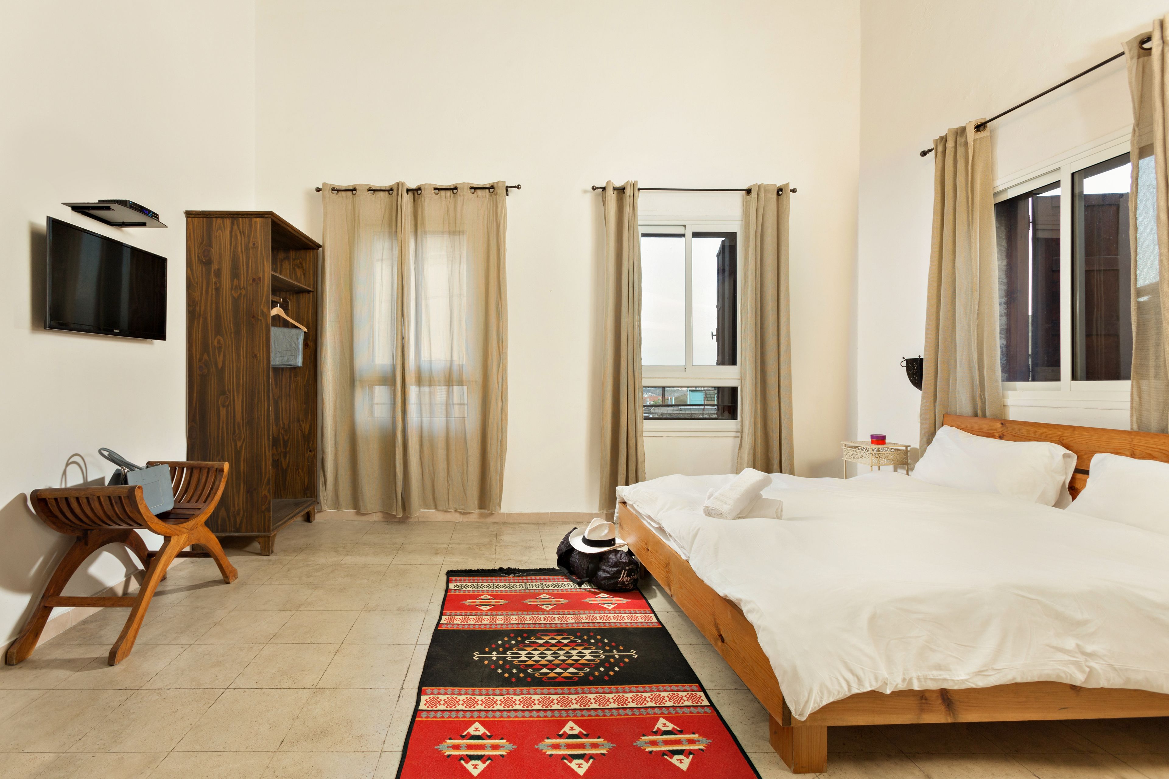 A hotel room with a bed, a TV mounted on the wall, a wardrobe, a desk with a chair, and a window with curtains.