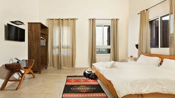 A hotel room with a bed, a TV mounted on the wall, a wardrobe, a desk with a chair, and a window with curtains.