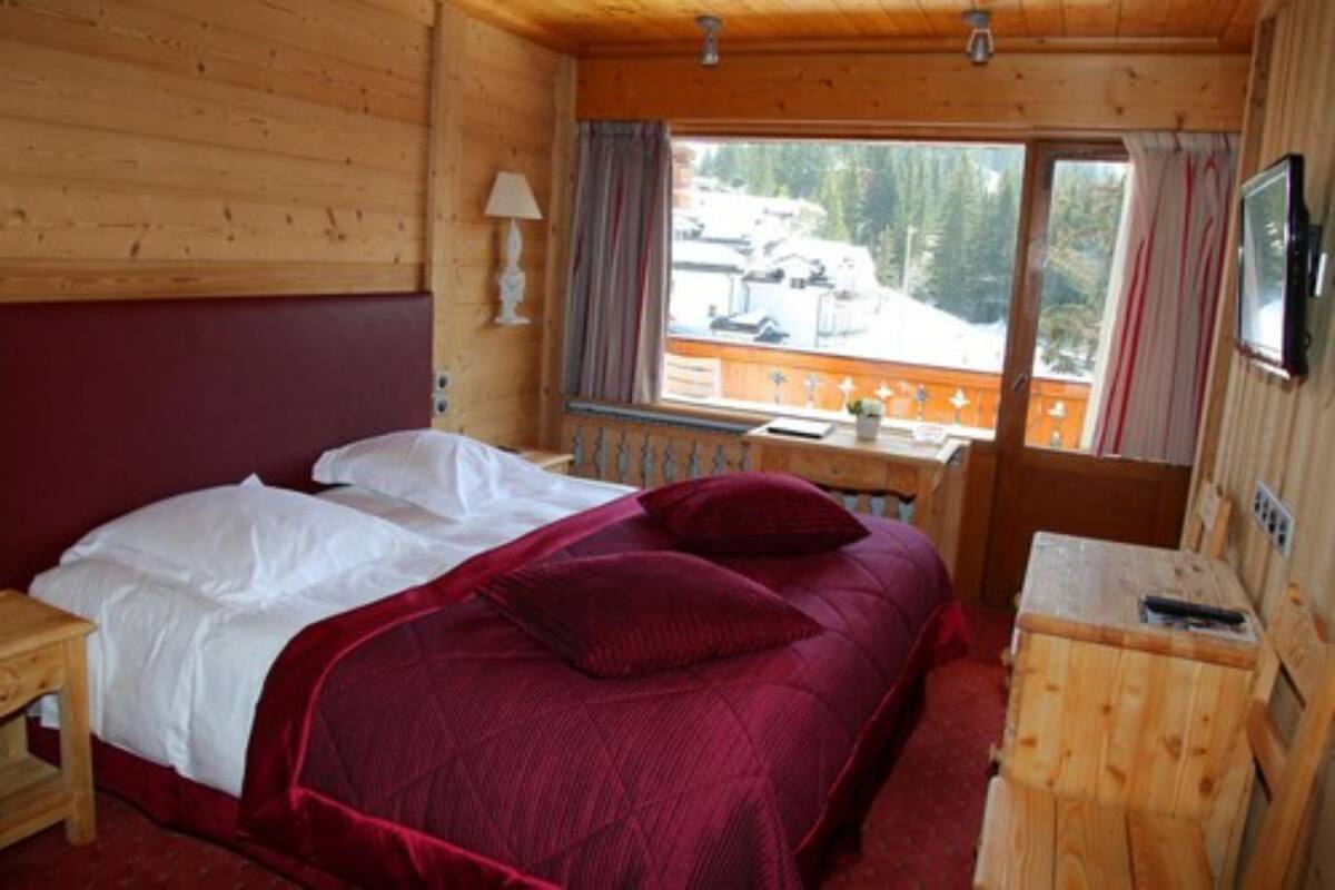 Une chambre dans un chalet en bois, avec un lit, un petit bureau, une fenêtre avec des rideaux et une vue sur un paysage enneigé.