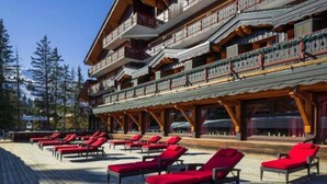 A multi-story building with balconies, a wooden deck with red lounge chairs, and a mountainous backdrop.