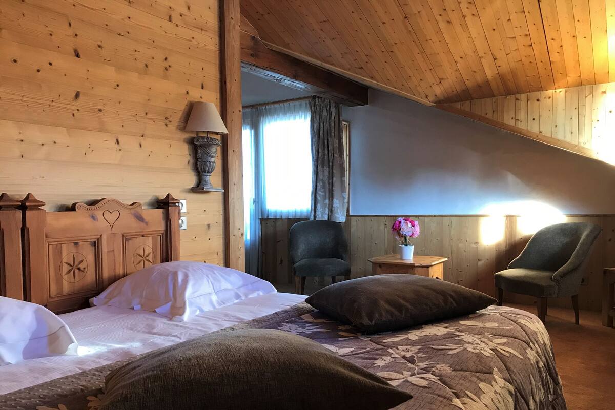 Une pièce chaleureuse aux murs en bois, avec un lit recouvert d’une courtepointe à motifs floraux, deux chaises et une petite table sur laquelle se trouve une lampe.