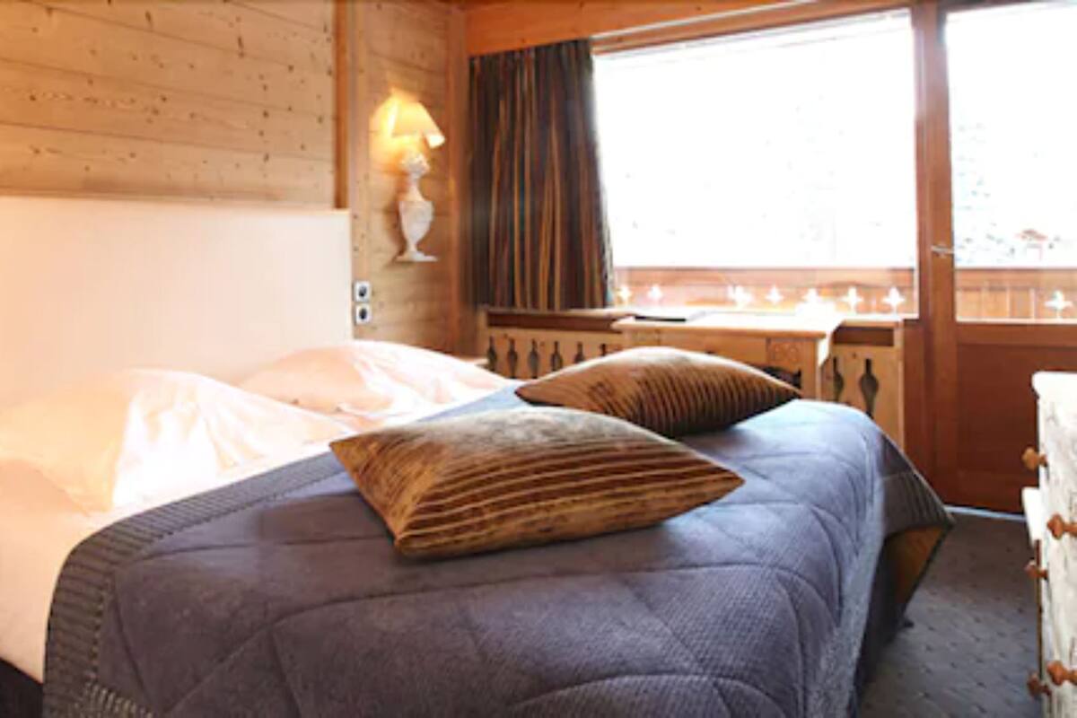 Une chambre à coucher avec un lit, des oreillers, une table de chevet, une lampe et une fenêtre avec des rideaux.