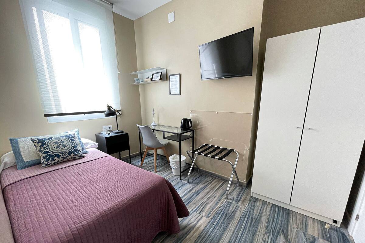 A hotel room with a bed, desk, chair, TV, wardrobe, and a window with blinds.