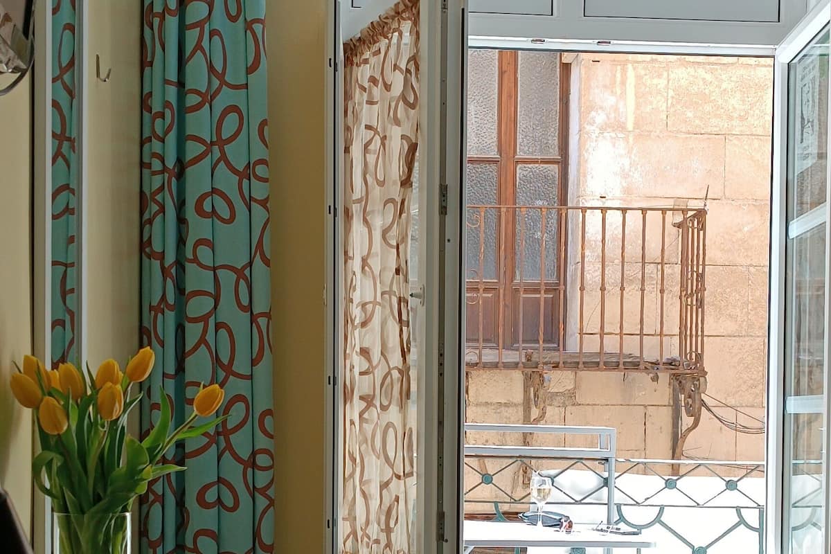 A room with a glass table, a chair, a bed with patterned bedding, a vase with flowers, and a view of the outside balcony.