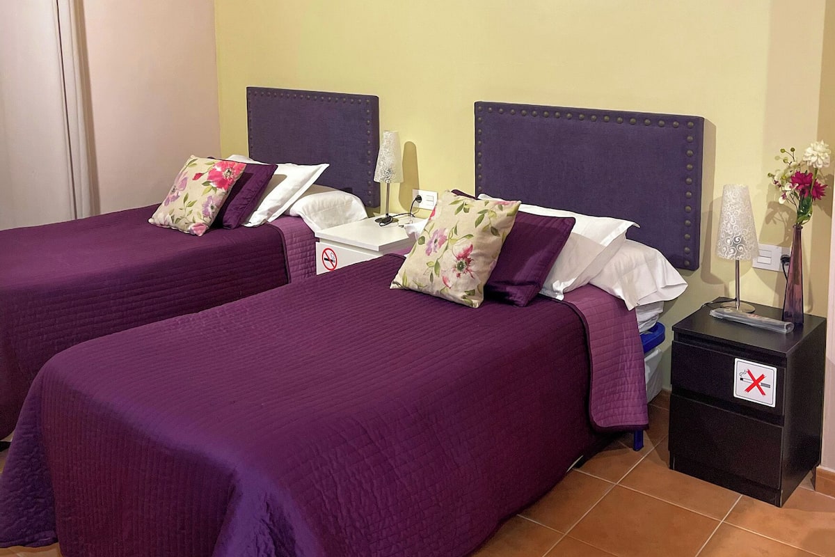 A hotel room with two beds, each with purple bedding and floral pillows, a nightstand with a lamp, and a vase with flowers.