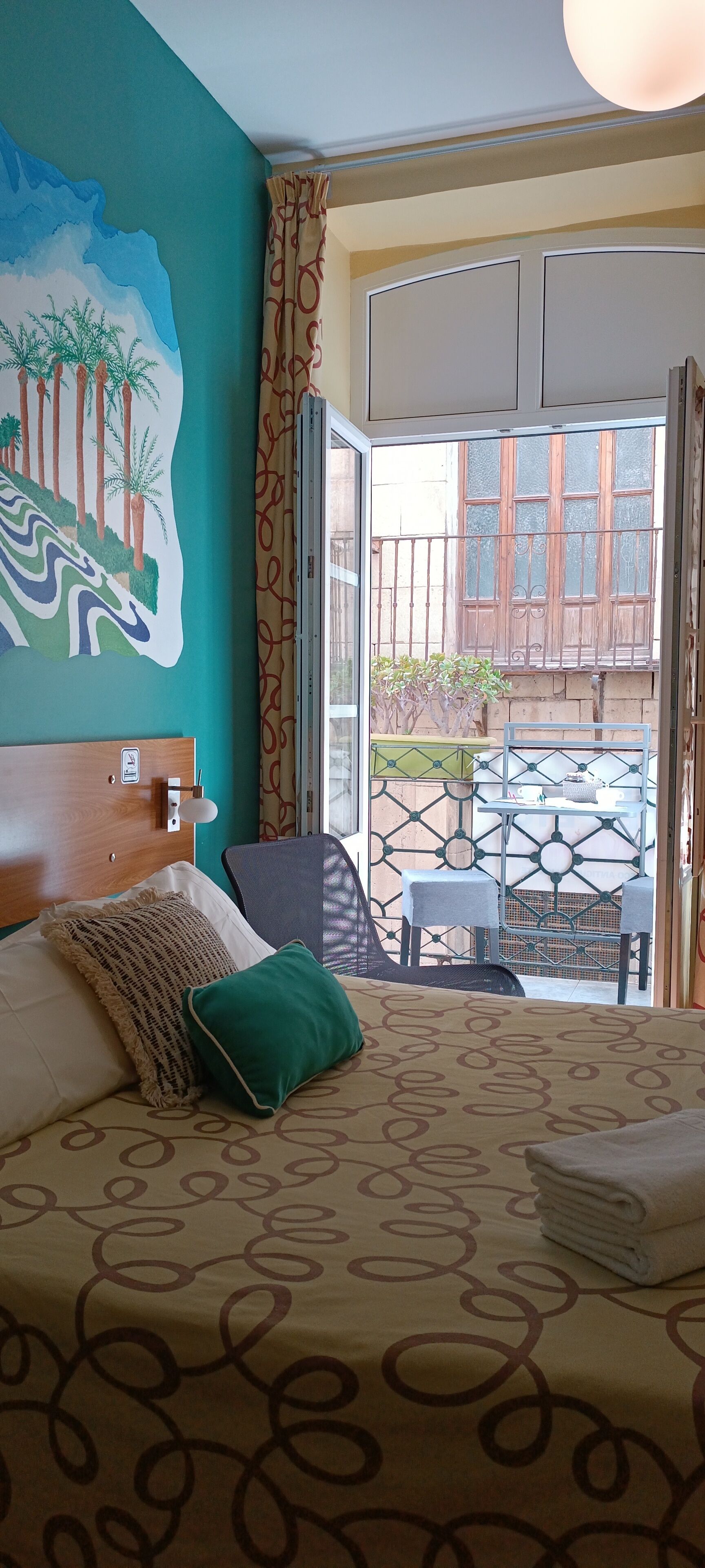 A hotel room with a bed, a painting of a landscape, a balcony with a table and chairs, and a view of the outside.