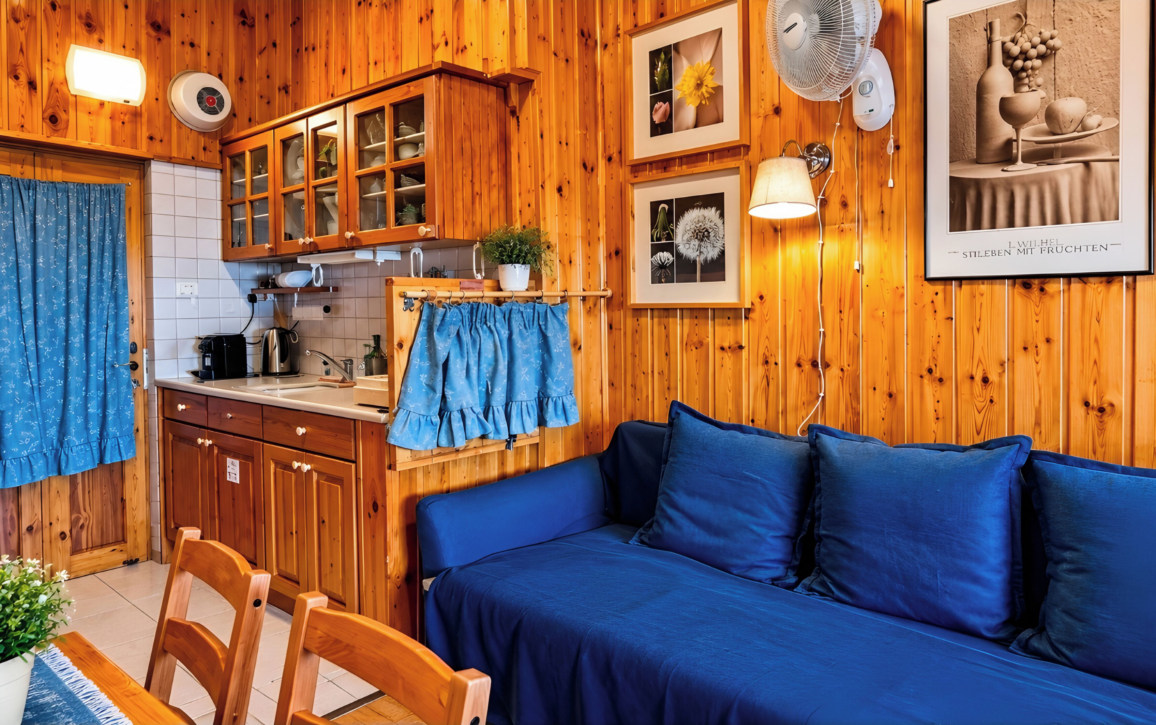 A cozy room with wooden walls, a blue sofa, a wooden dining table, and a kitchen area with cabinets and a kettle.