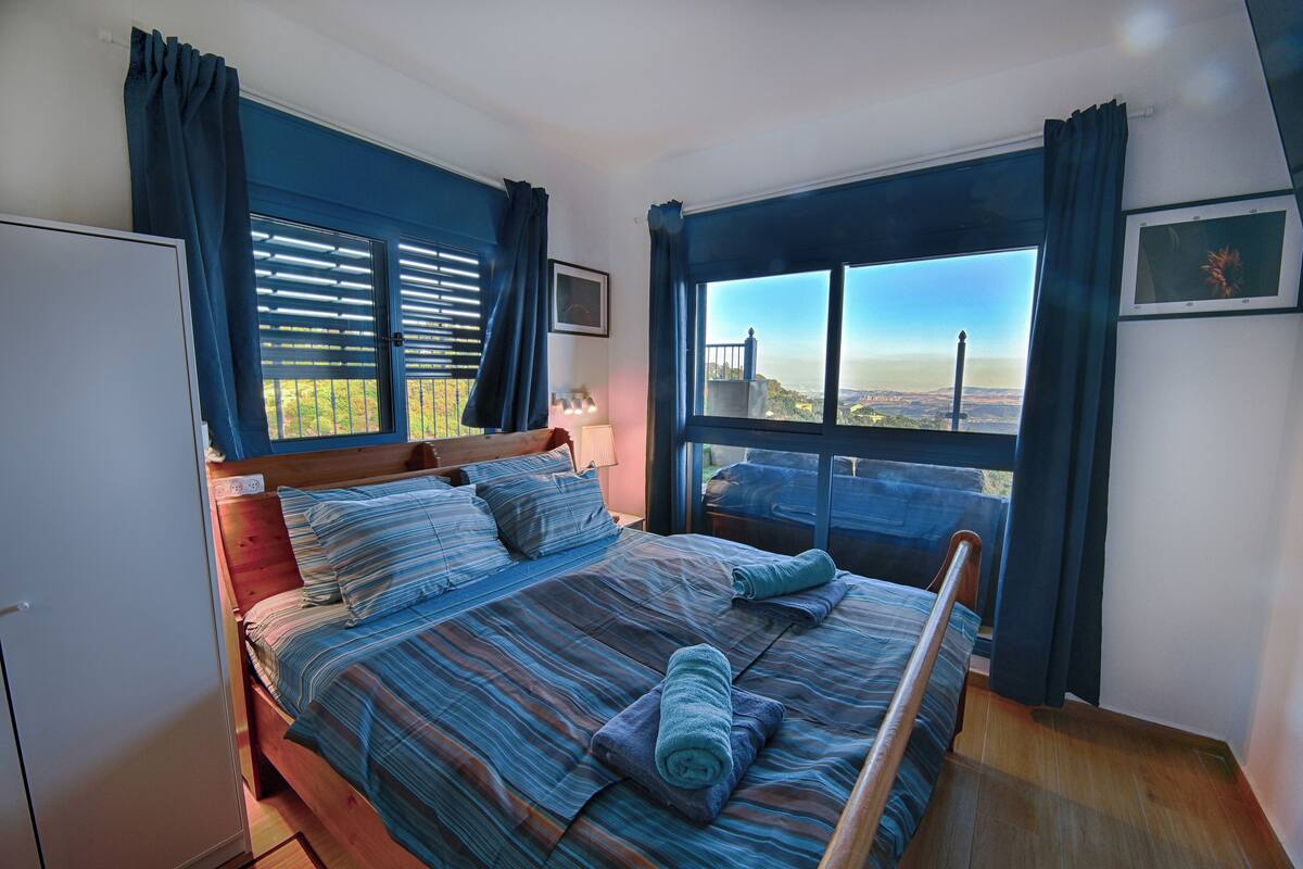 A bedroom with a large bed, a view of the cityscape, and a window with blinds.