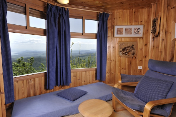 A wooden cabin room with a bed, two blue chairs, a round table, and a view of mountains through large windows.