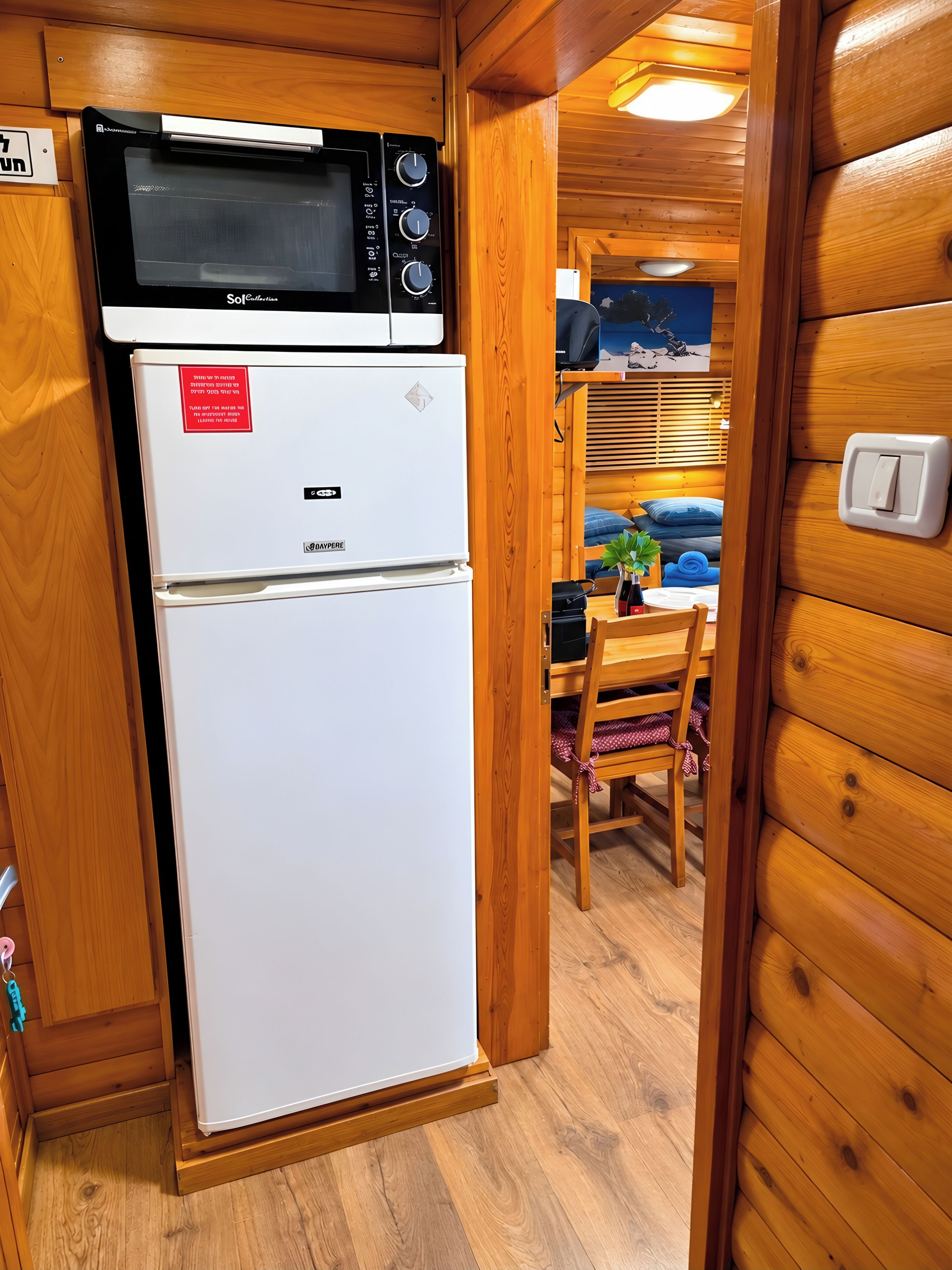 A compact kitchen with a microwave and refrigerator, wooden walls, a dining area with a table and chairs, and a visible light switch.