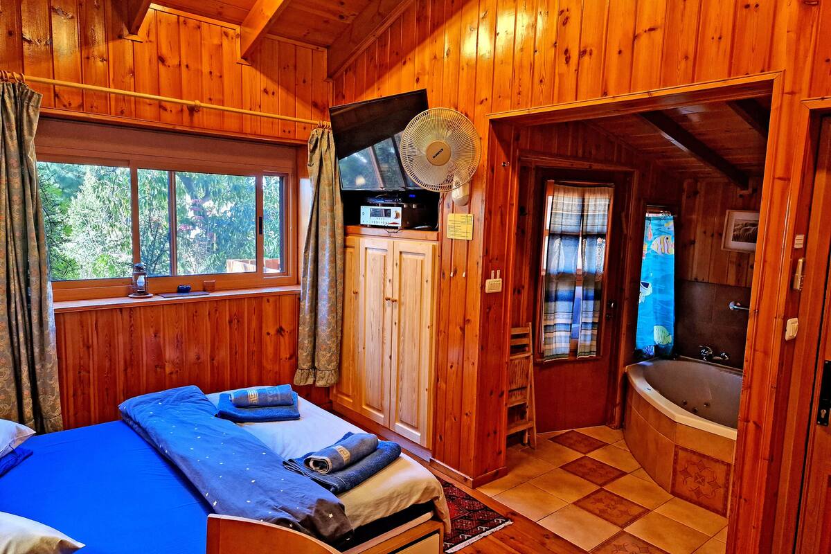 A wooden cabin bedroom with a bed, a fan, a window, and a bathroom with a bathtub.
