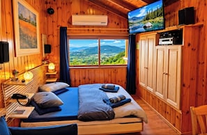 A wooden room with a bed, a television, and a view of a mountainous landscape. - Ohn-Bar Guesthouse (Merom HaGalil)