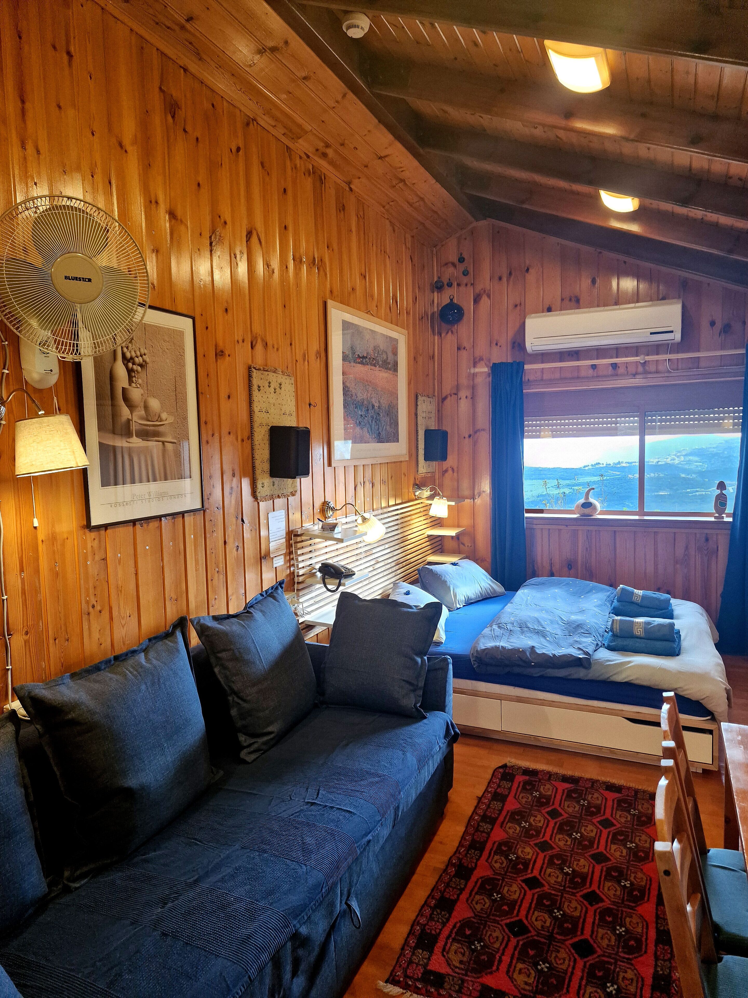 A wooden cabin room with a sofa, two beds, a window with a view, and a ceiling fan.