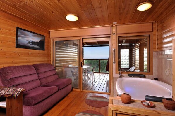 A wooden cabin interior with a sofa, a bathtub, and a sliding glass door leading to an outdoor deck.