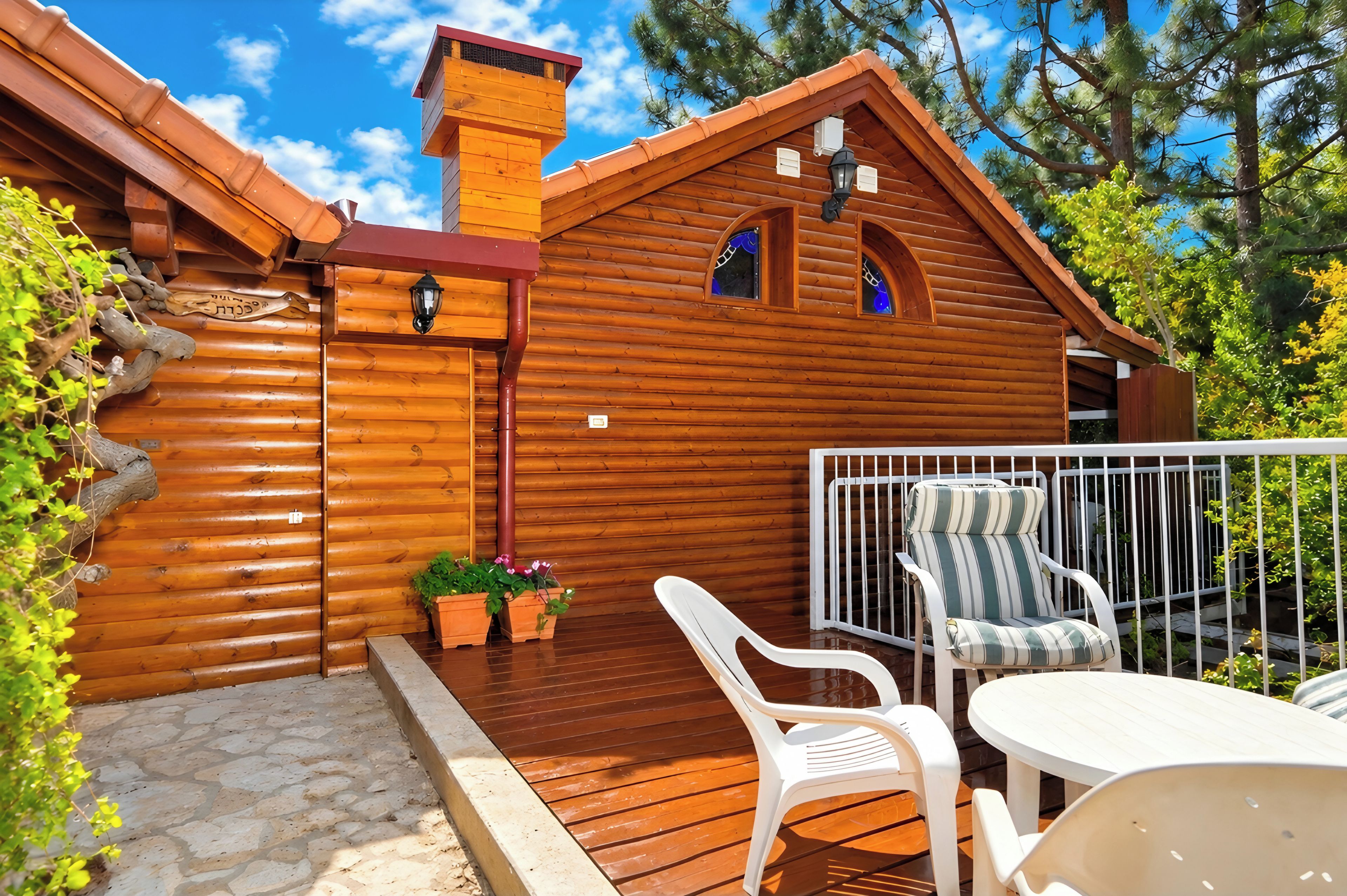 A wooden cabin with a balcony, white plastic chairs, and potted plants.