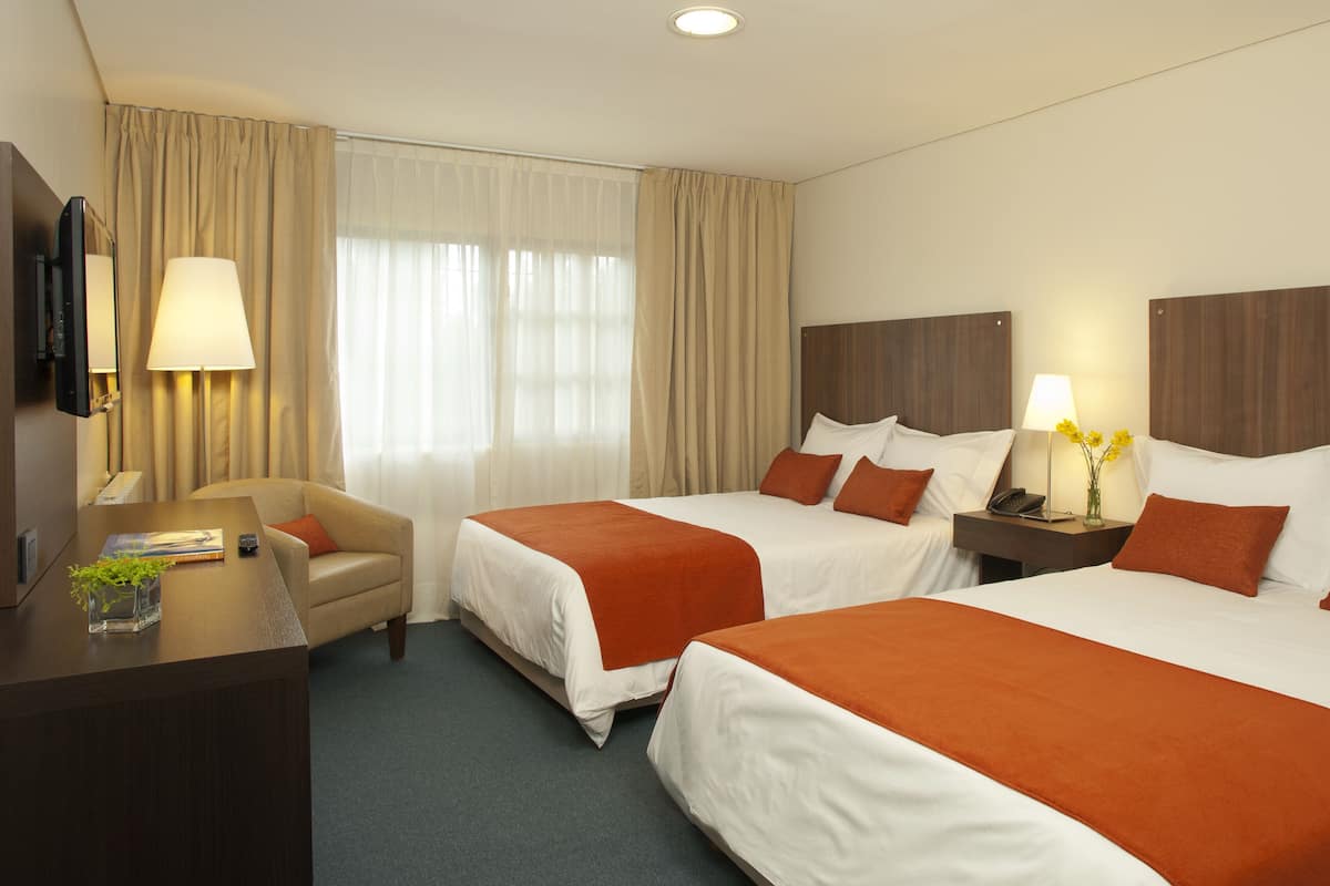 Une chambre d’hôtel avec deux lits, un bureau, une chaise, une lampe et une fenêtre avec des rideaux.