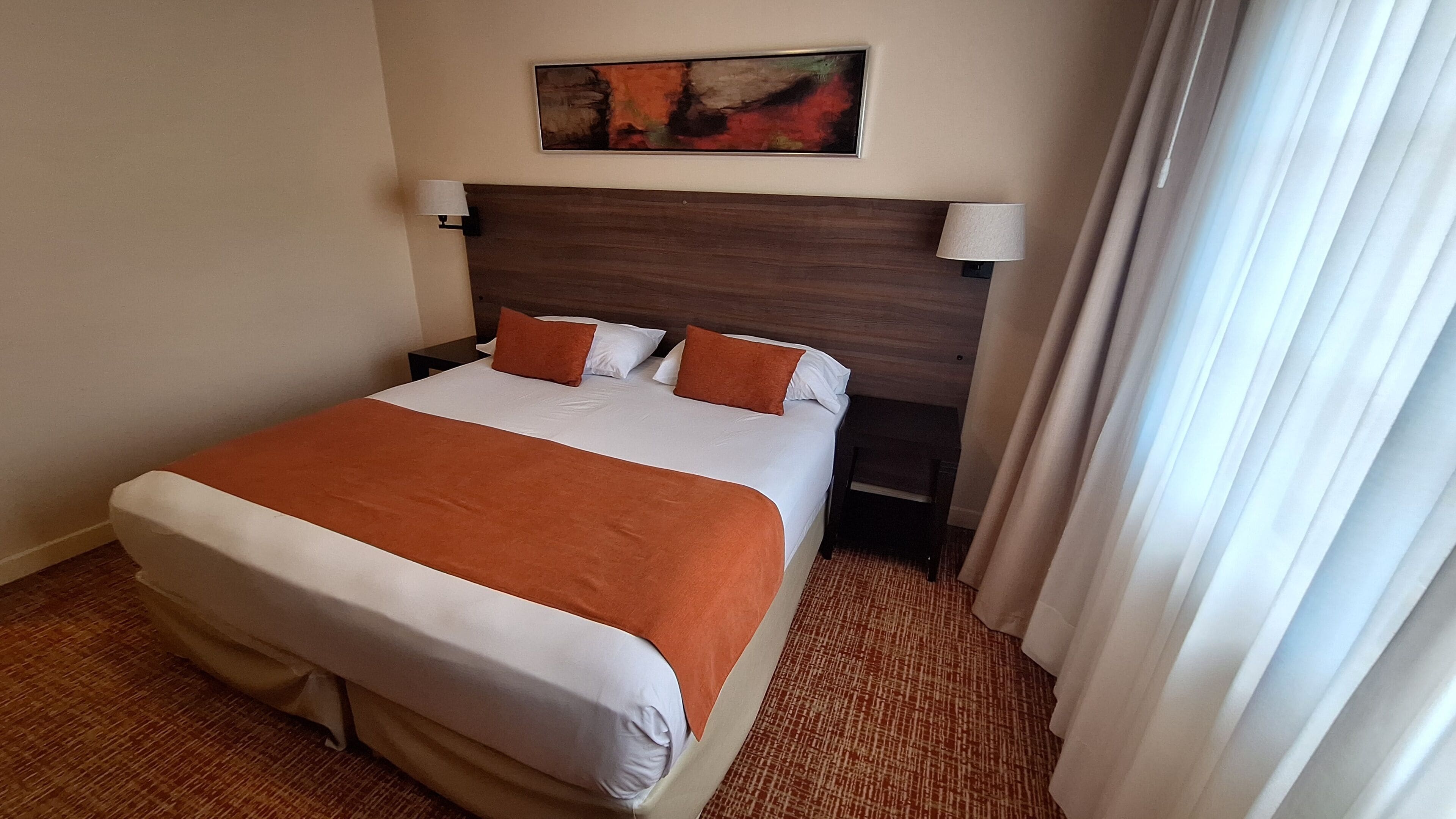 A hotel room with a large bed, two bedside tables, a headboard, a painting on the wall, and a window with curtains.